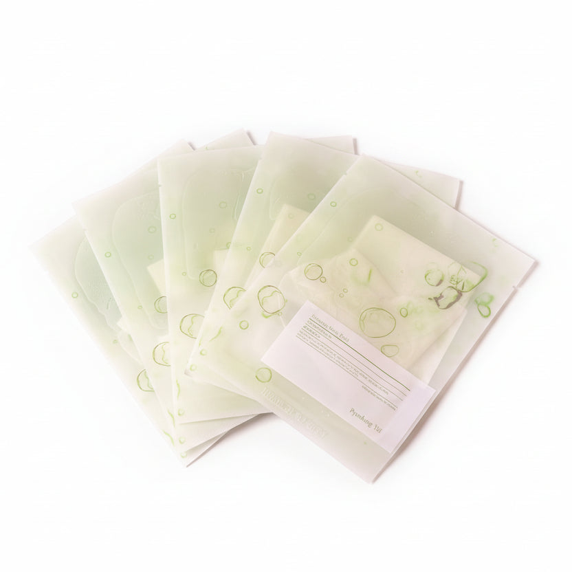 Stack of green and white packets on a wooden surface with a pen and colorful object in the background.